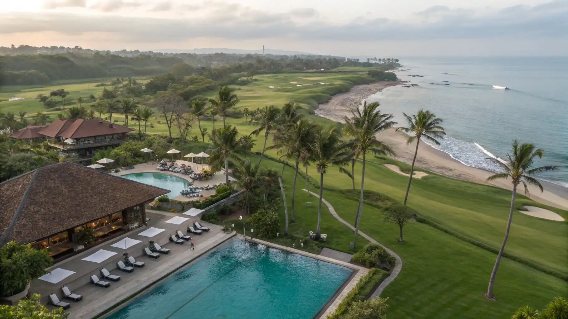 Aerial view of a coastal resort with pools and lawns running down to the beach